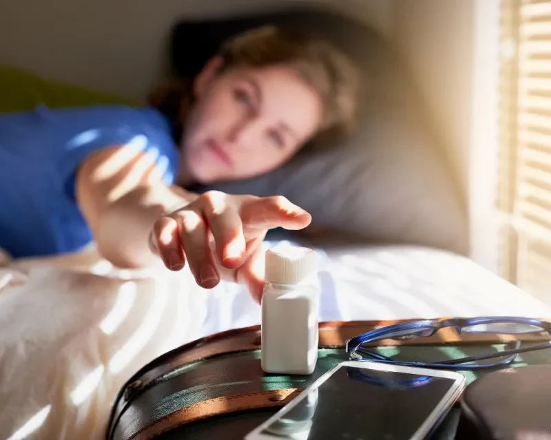 A woman reaching for a bottle of vitamins for women on a bedside table, highlighting the importance of daily supplementation for health.