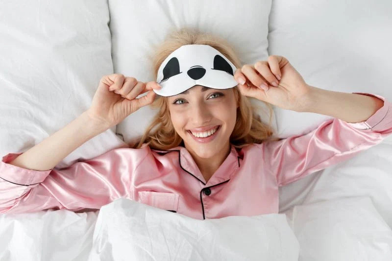 A woman smiling in bed while removing her panda sleep mask, ready for a restful night with the help of sleep supplements