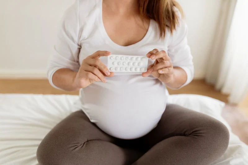 A pregnant woman holding a pack of prenatal supplements, sitting on the bed and preparing to take her daily vitamins for a healthy pregnancy